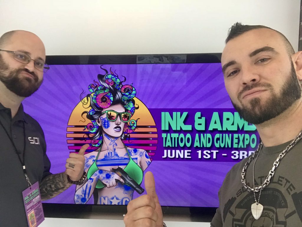 Two men pointing to a sign that says Ink & Arms