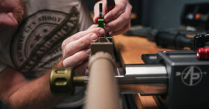 Close-up of a gunsmith working on a rifle barrel using a barrel vise and tools in a workshop.